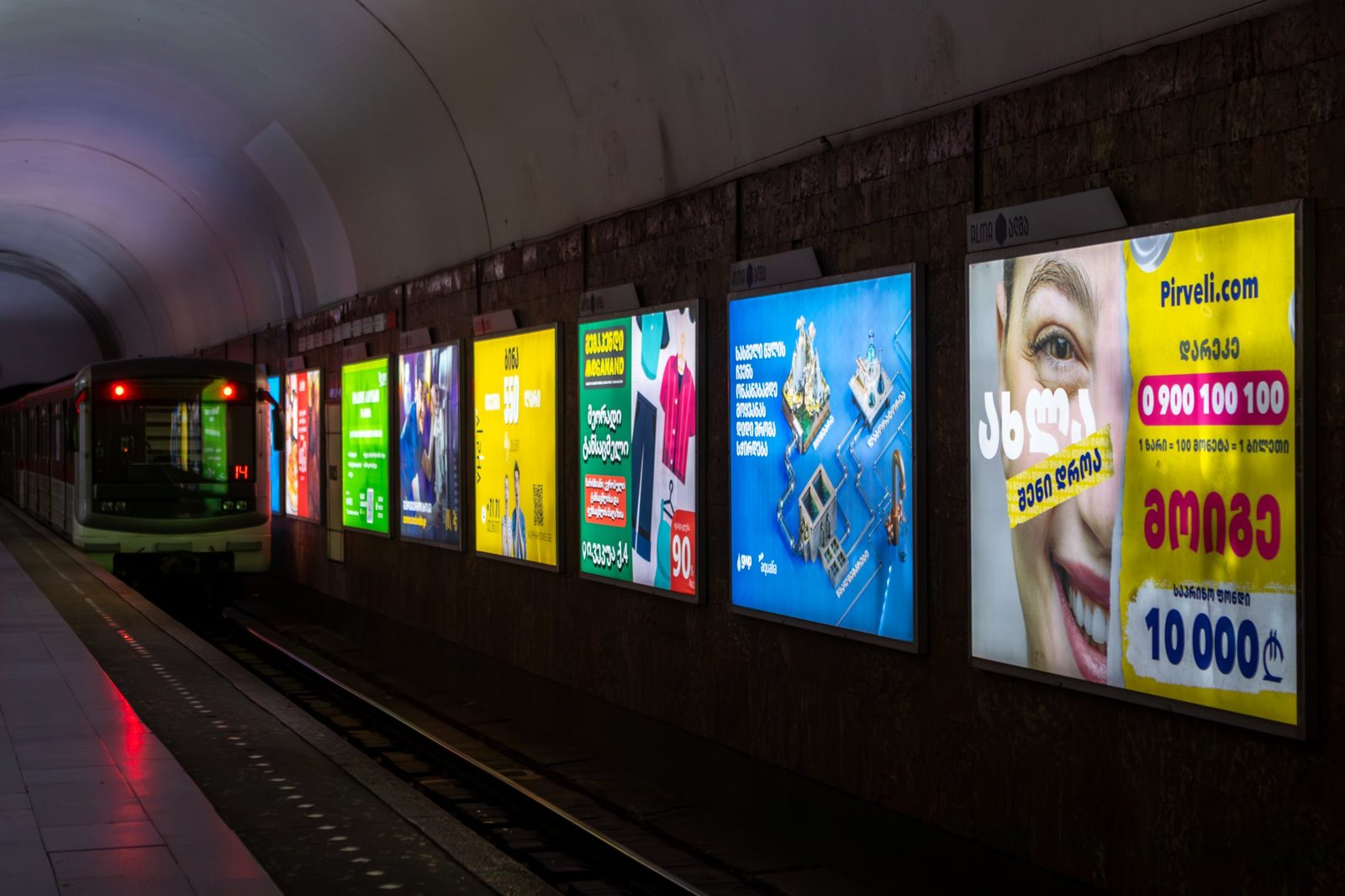 A row of ALMA backlit panels along a Tbilisi metro platform as a train arrives